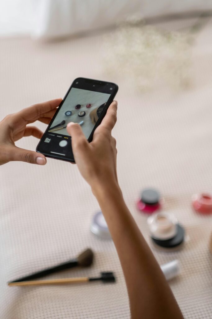 A person using a smartphone to capture an image of various makeup products on a bed.