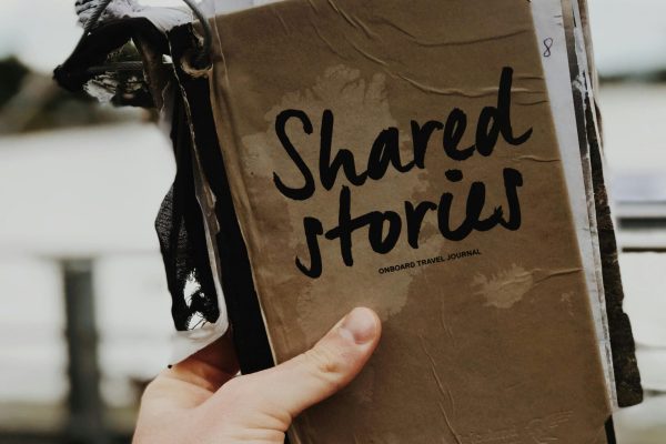 Hand holds a "Shared Stories" journal against a blurred outdoor backdrop, emphasizing creativity and personal storytelling.